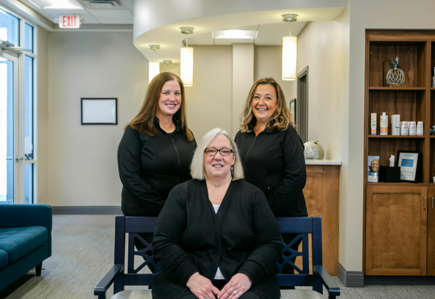 Three administrators from Progressive Dental Care, smiling and dressed in black, standing and sitting in a welcoming office environment with modern decor and dental products displayed in the background.